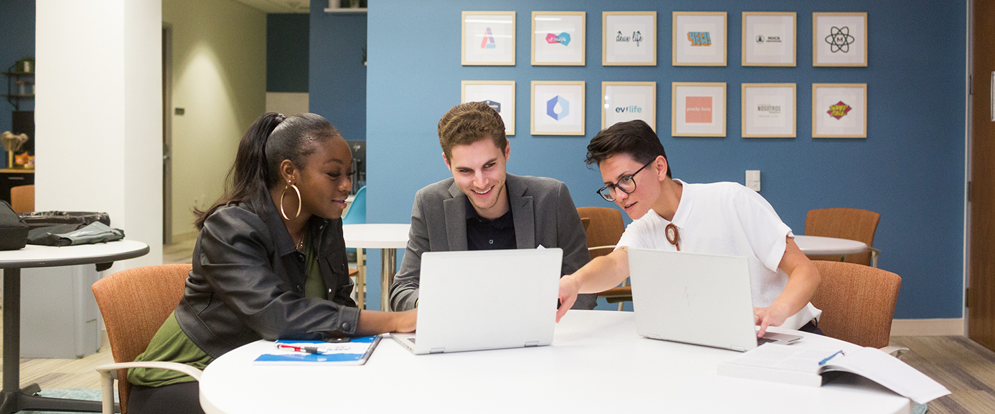 three students working together at a workstation, two laptops are open